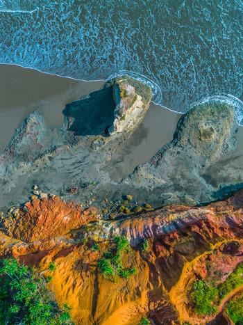 Aerial view of rugged coastal cliffs with vibrant orange and red soil unique rock formations on the sandy beach and turquoise ocean waves crashing against the shore in Bali Bali Indonesia. Aerial view of rugged coastal cliffs with vibrant orange and red soil unique rock formations on the sandy beach and turquoise ocean waves crashing against the shore in Bali Bali Indonesia.