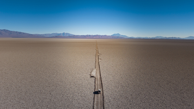 Aerial view of a lone vehicle driving along a straight dirt road through the vast arid salt flats of Tolar Grande Salta Province Argentina. Aerial view of a lone vehicle driving along a straight dirt road through the vast arid salt flats of Tolar Grande Salta Province Argentina.