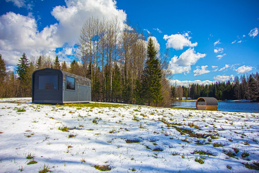 View of a modern dark cabin and a barrel sauna situated in a snowy field beside a lake surrounded by a forest under a bright blue sky in Latvia. View of a modern dark cabin and a barrel sauna situated in a snowy field beside a lake surrounded by a forest under a bright blue sky in Latvia.