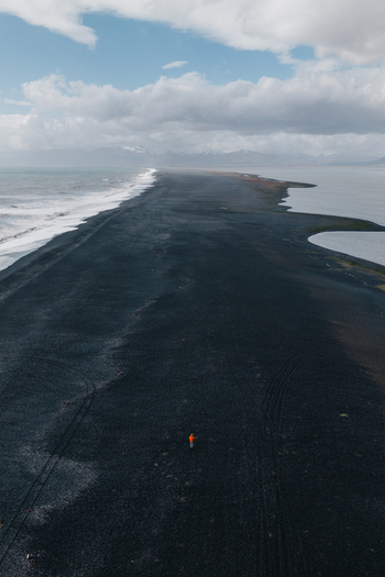Aerial view of the black sand beach at Stokksnes with a person in an orange jacket walking on the dark volcanic shore surrounded by the ocean and distant mountains in Iceland.