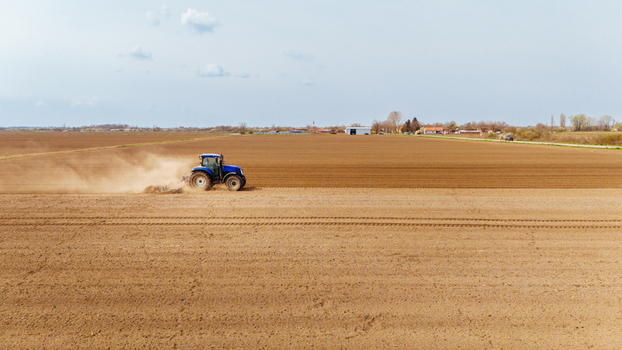 Aerial view of a blue tractor working the light brown soil of a vast field kicking up dust under a pale sky Sremska Mitrovica Vojvodina Serbia. Aerial view of a blue tractor working the light brown soil of a vast field kicking up dust under a pale sky Sremska Mitrovica Vojvodina Serbia.