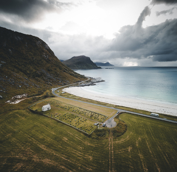Aerial view of Vik Strand beach with a small cemetery and church nearby coastal road mountains and sea under a cloudy sky in Leknes Nordland Norway.