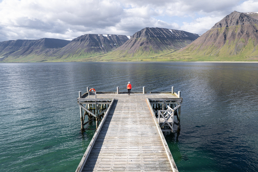 Aerial view of Onundarfjordur Pier with a person in an orange jacket looking out over the calm fjord towards massive flat topped mountains under a cloudy sky in Holt Isafjardabaer Iceland. Aerial view of Onundarfjordur Pier with a person in an orange jacket looking out over the calm fjord towards massive flat topped mountains under a cloudy sky in Holt Isafjardabaer Iceland.