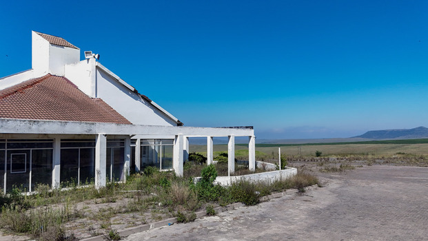 Aerial view of an abandoned white building with a red tiled roof and overgrown vegetation overlooking a vast open landscape under a clear blue sky in iNtabazwe Free State South Africa. Aerial view of an abandoned white building with a red tiled roof and overgrown vegetation overlooking a vast open landscape under a clear blue sky in iNtabazwe Free State South Africa.