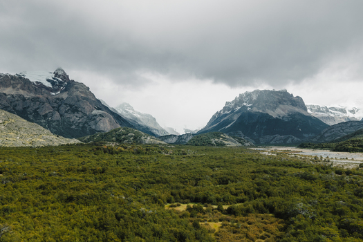 Aerial view of the rugged peaks of El Chalten rising above lush green forest and a winding riverbed under a dramatic cloudy sky in El Chalten Santa Cruz Province Argentina. Aerial view of the rugged peaks of El Chalten rising above lush green forest and a winding riverbed under a dramatic cloudy sky in El Chalten Santa Cruz Province Argentina.