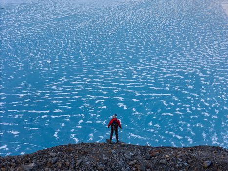 Aerial view of a hiker in a red jacket standing on a rocky ridge overlooking a vast frozen glacial lake with intricate white ice patterns in Nepal.