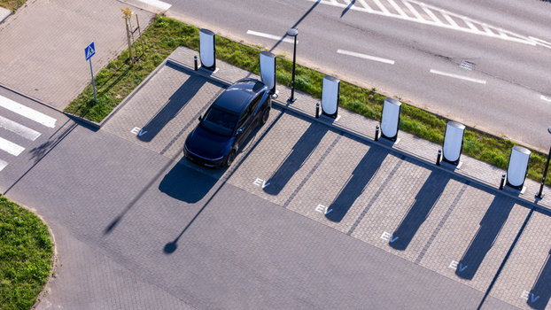 Aerial view of an electric vehicle charging station with a dark car parked at a charging point featuring paved parking spaces with EV markings and a nearby road in Gdansk Poland. Aerial view of an electric vehicle charging station with a dark car parked at a charging point featuring paved parking spaces with EV markings and a nearby road in Gdansk Poland.