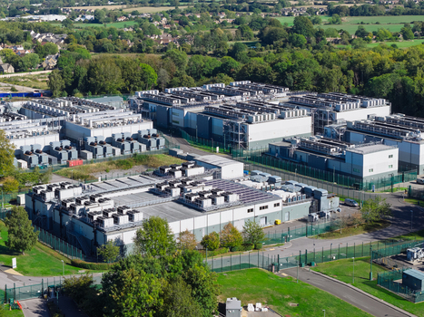 Aerial view of a large data center facility featuring multiple industrial buildings with rooftop cooling units surrounded by security fencing and lush green landscape in the United Kingdom.