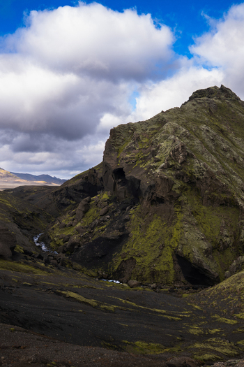 View of rugged moss covered mountains rise dramatically against a backdrop of a cloudy blue sky a river snaking through the valley floor Skaftárhreppur Iceland. View of rugged moss covered mountains rise dramatically against a backdrop of a cloudy blue sky a river snaking through the valley floor Skaftárhreppur Iceland.