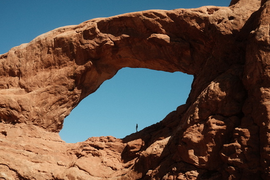 View of Arches National Park with a person standing inside a massive natural sandstone arch under a clear blue sky in Grand County Utah United States. View of Arches National Park with a person standing inside a massive natural sandstone arch under a clear blue sky in Grand County Utah United States.