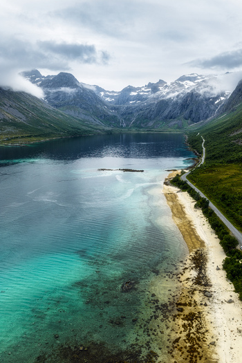 Aerial view of Kvaloya featuring turquoise coastal waters a winding road along the shoreline and dramatic snow capped mountains in Tromso Municipality Troms Norway. Aerial view of Kvaloya featuring turquoise coastal waters a winding road along the shoreline and dramatic snow capped mountains in Tromso Municipality Troms Norway.