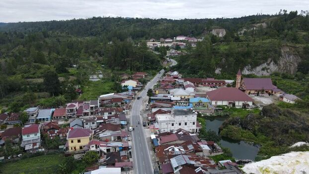 Aerial view of the town of Tarutung featuring a central road residential houses with red roofs and a prominent church building surrounded by lush green hills and vegetation in Tarutung North Sumatra Indonesia. Aerial view of the town of Tarutung featuring a central road residential houses with red roofs and a prominent church building surrounded by lush green hills and vegetation in Tarutung North Sumatra Indonesia.