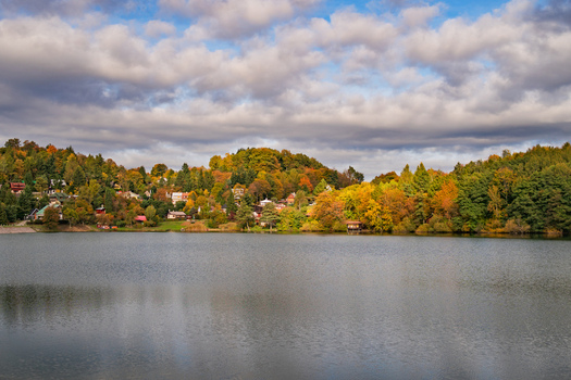 View of Velka Richnava lake with colorful autumn trees and hillside houses under a cloudy sky in Stiavnicke Bane Banska Bystrica Regio Slovakia.