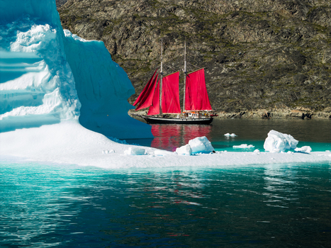Aerial view of a sailboat with vibrant red sails navigating near a massive blue iceberg in the turquoise waters of a rocky Greenland. Aerial view of a sailboat with vibrant red sails navigating near a massive blue iceberg in the turquoise waters of a rocky Greenland.