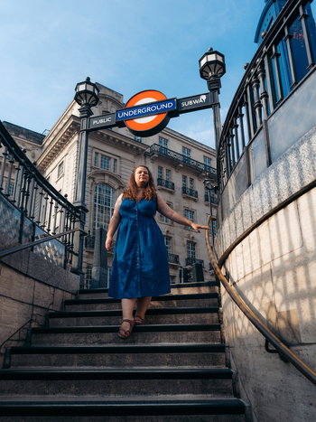 View of a woman ascending sunlit stone steps from the subterranean depths her vibrant blue dress contrasting with the iconic Underground sign and grand architecture London England United Kingdom. View of a woman ascending sunlit stone steps from the subterranean depths her vibrant blue dress contrasting with the iconic Underground sign and grand architecture London England United Kingdom.