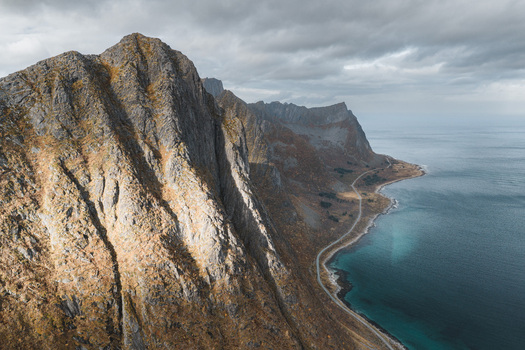 Aerial view of Senja island featuring dramatic rugged mountain peaks a winding coastal road and turquoise sea under a cloudy sky in Senja Troms Norway.