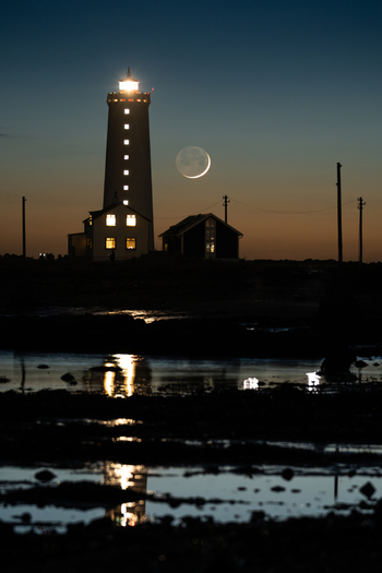 View of Grotta Lighthouse illuminated at twilight with a crescent moon in the sky and reflections on the water in Seltjarnarnes Seltjarnarnesbaer Iceland. View of Grotta Lighthouse illuminated at twilight with a crescent moon in the sky and reflections on the water in Seltjarnarnes Seltjarnarnesbaer Iceland.