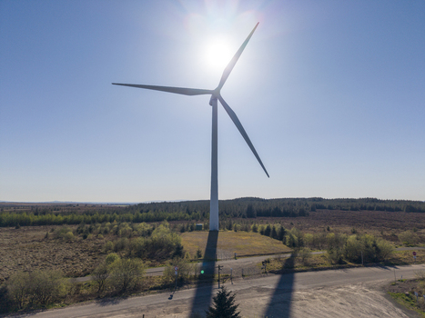 Aerial view of a tall white wind turbine standing in a vast rural landscape with coniferous forests and a paved road under a brilliant sun and clear blue sky.