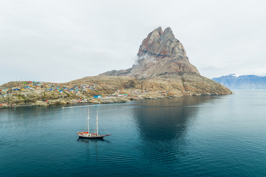 Aerial view of the iconic Uummannaq mountain rising above the colorful houses of the coastal town and a sailboat gliding through the calm dark blue waters in Uummannaq Kitaa Greenland. Aerial view of the iconic Uummannaq mountain rising above the colorful houses of the coastal town and a sailboat gliding through the calm dark blue waters in Uummannaq Kitaa Greenland.