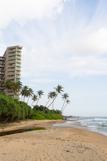 View of a sandy beach with leaning palm trees and a modern high rise building under a bright sky in Galle Southern Province Sri Lanka.