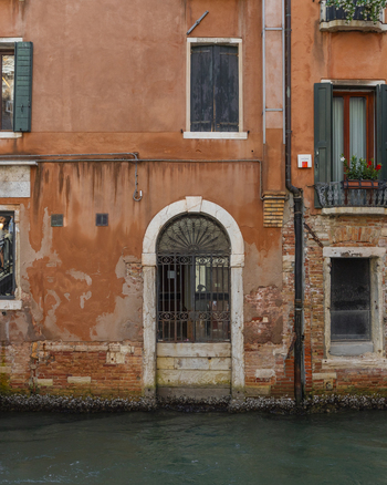 View of a weathered orange building facade with an arched doorway and iron gate along a canal in Venice Veneto Italy. View of a weathered orange building facade with an arched doorway and iron gate along a canal in Venice Veneto Italy.