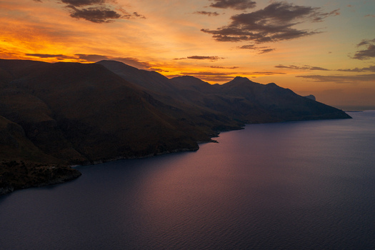 Aerial view of the rugged coastline of Riserva Naturale Orientata dello Zingaro at sunset with mountains meeting the sea under a golden sky in San Vito Lo Capo Sicilia Italy. Aerial view of the rugged coastline of Riserva Naturale Orientata dello Zingaro at sunset with mountains meeting the sea under a golden sky in San Vito Lo Capo Sicilia Italy.