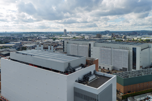 Aerial view of a large modern data center complex with industrial rooftops and ventilation systems set against an urban landscape under a cloudy sky in the United Kingdom. Aerial view of a large modern data center complex with industrial rooftops and ventilation systems set against an urban landscape under a cloudy sky in the United Kingdom.