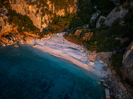 Aerial view of Spiaggia di Cala Fuili pebble beach nestled between steep limestone cliffs and lush greenery at sunset in Dorgali Sardegna Italy. Aerial view of Spiaggia di Cala Fuili pebble beach nestled between steep limestone cliffs and lush greenery at sunset in Dorgali Sardegna Italy.