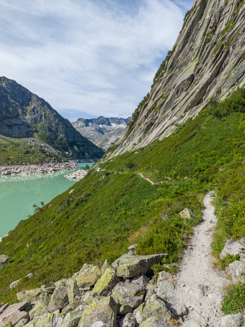 Aerial view of Gelmersee alpine reservoir with turquoise water steep rocky slopes and a hiking trail winding through green mountain vegetation in Switzerland.