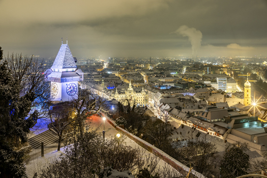 View of the illuminated clock tower standing tall amidst the snowy landscape with city lights twinkling below in Graz Styria Austria. View of the illuminated clock tower standing tall amidst the snowy landscape with city lights twinkling below in Graz Styria Austria.