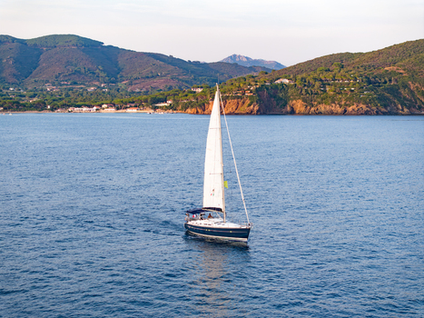 Aerial view of a sailboat sailing in the blue waters of Lacona Bay with lush green hills and coastal cliffs in the background in Lacona Tuscany Italy.