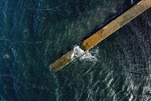 Aerial view of a concrete pier extending into the dark choppy sea with waves crashing against the structure in Stein Scotland United Kingdom. Aerial view of a concrete pier extending into the dark choppy sea with waves crashing against the structure in Stein Scotland United Kingdom.