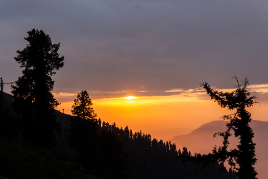 View of the sun sinking behind silhouetted trees on a mountain ridge with a warm glow illuminating the sky Pir Chanasi Azad Kashmir Pakistan. View of the sun sinking behind silhouetted trees on a mountain ridge with a warm glow illuminating the sky Pir Chanasi Azad Kashmir Pakistan.