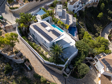 Aerial view of a modern white villa featuring rooftop solar panels a private swimming pool and palm trees in a hillside residential area in Moraira Valencian Community Spain.
