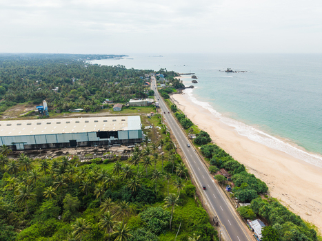 Aerial view of the coastal road running alongside a sandy beach and dense palm trees near an industrial building in Peraliya Southern Province Sri Lanka. Aerial view of the coastal road running alongside a sandy beach and dense palm trees near an industrial building in Peraliya Southern Province Sri Lanka.