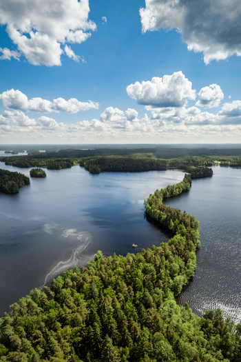 Aerial view of Liesjarvi National Park featuring a winding forested esker cutting through the deep blue lake waters under a bright sky with fluffy clouds in Tammela Finland. Aerial view of Liesjarvi National Park featuring a winding forested esker cutting through the deep blue lake waters under a bright sky with fluffy clouds in Tammela Finland.