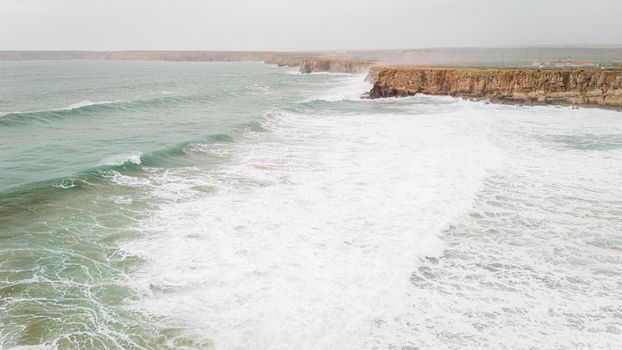 Aerial view of the rugged cliffs of Sagres with powerful white waves crashing against the rocky coastline under a misty sky in Sagres Faro Portugal. Aerial view of the rugged cliffs of Sagres with powerful white waves crashing against the rocky coastline under a misty sky in Sagres Faro Portugal.
