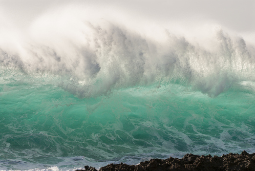 View of a powerful turquoise ocean wave breaking with white sea spray over dark volcanic rocks on the North Shore in Haleiwa Hawaii United States.