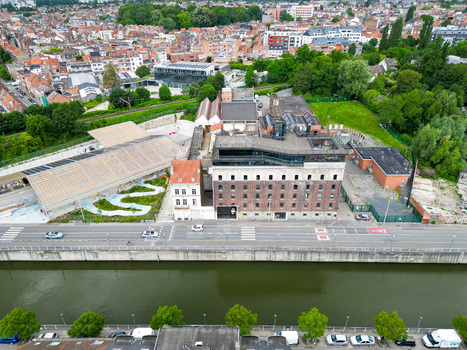 Aerial view of a large brick industrial building with a modern glass rooftop extension and a wavy garden path next to a canal in Anderlecht Brussels Belgium.