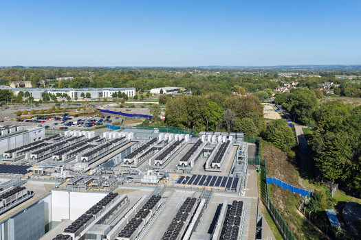 Aerial view of a large data center facility featuring rows of cooling units on the roof surrounded by lush green trees and a parking area in the United Kingdom.