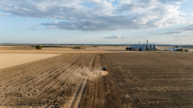 Aerial view of a tractor plowing a large agricultural field creating a dust trail with grain silos in the background under a cloudy sky in Veliki Radinci Vojvodina Serbia. Aerial view of a tractor plowing a large agricultural field creating a dust trail with grain silos in the background under a cloudy sky in Veliki Radinci Vojvodina Serbia.