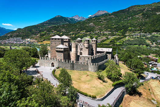 Aerial view of the medieval Fenis Castle with its stone towers and crenelated walls surrounded by green hills and Alpine mountains in Fenis Aosta Valley Italy. Aerial view of the medieval Fenis Castle with its stone towers and crenelated walls surrounded by green hills and Alpine mountains in Fenis Aosta Valley Italy.