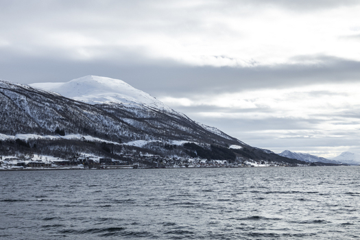 View of snow covered mountains overlooking a coastal town and the dark waters of a fjord under a cloudy winter sky Tromso Troms Norway.