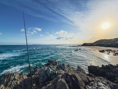 View of a fishing rod on a rocky coastline with waves crashing and a sandy beach under a bright sun in Plettenberg Bay Western Cape South Africa. View of a fishing rod on a rocky coastline with waves crashing and a sandy beach under a bright sun in Plettenberg Bay Western Cape South Africa.