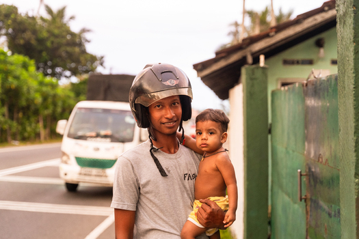 Sri Lanka 14 January 2026 View of a man wearing a motorcycle helmet holding a young child by a roadside with a house and lush greenery in the background.