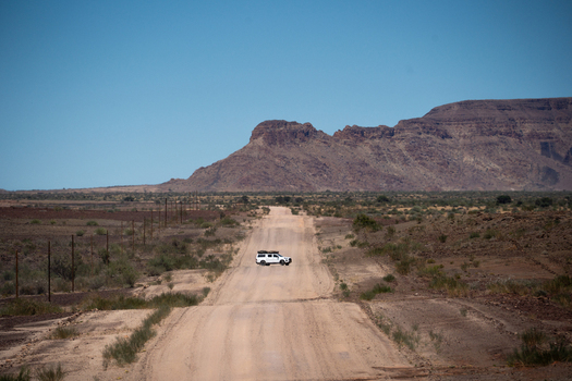 View of a white vehicle navigates a long dusty road cutting through the arid landscape under a clear sky with distant rocky hills Keetmanshoop Namibia. View of a white vehicle navigates a long dusty road cutting through the arid landscape under a clear sky with distant rocky hills Keetmanshoop Namibia.