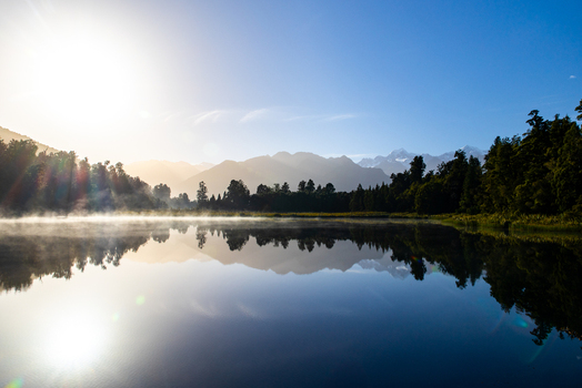 View of a serene lake mirroring the snow capped mountains under a radiant sun with mist rising gently over the still water Lake Matheson New Zealand. View of a serene lake mirroring the snow capped mountains under a radiant sun with mist rising gently over the still water Lake Matheson New Zealand.