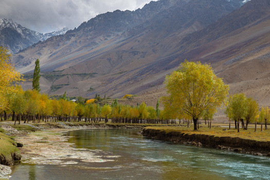 View of a tranquil river carving through a vibrant valley framed by golden autumn foliage and towering snow capped mountains beneath a dramatic sky Gilgit Pakistan.