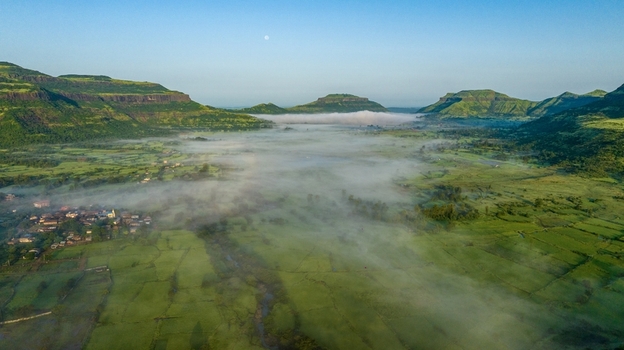 Aerial view of the lush green landscape of Igatpuri in the Western Ghats featuring rolling hills fields and morning mist under a clear blue sky Igatpuri Maharashtra India.