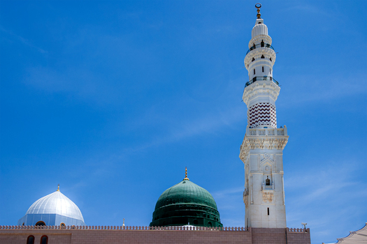 View of the iconic green dome and a tall white minaret of Al Masjid e Nabawi against a clear blue sky in Madinah Al Madinah Province Saudi Arabia.